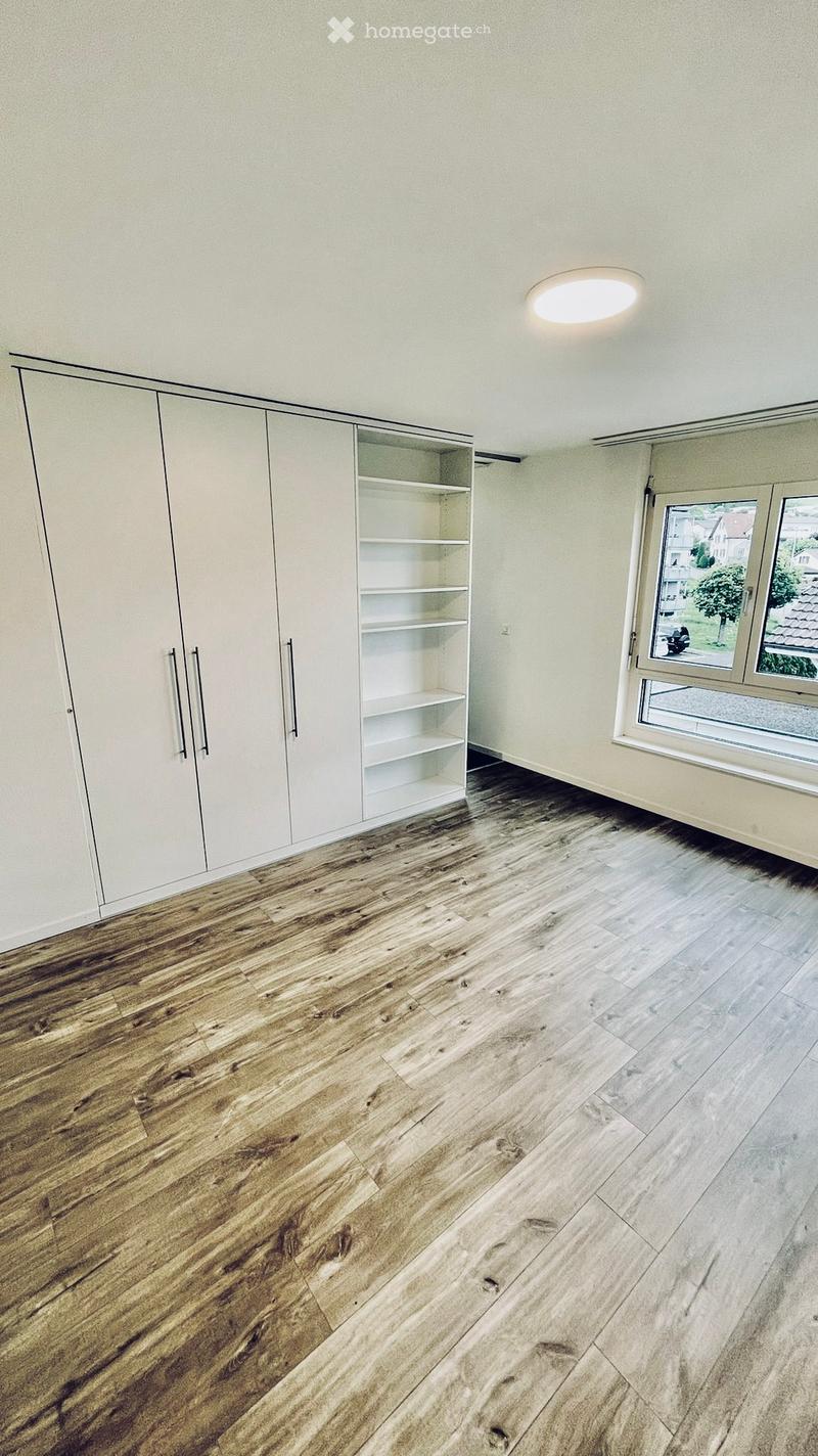 Empty room with light-colored walls, wooden flooring, built-in closet with empty shelves, and a large window with a view of the outside.