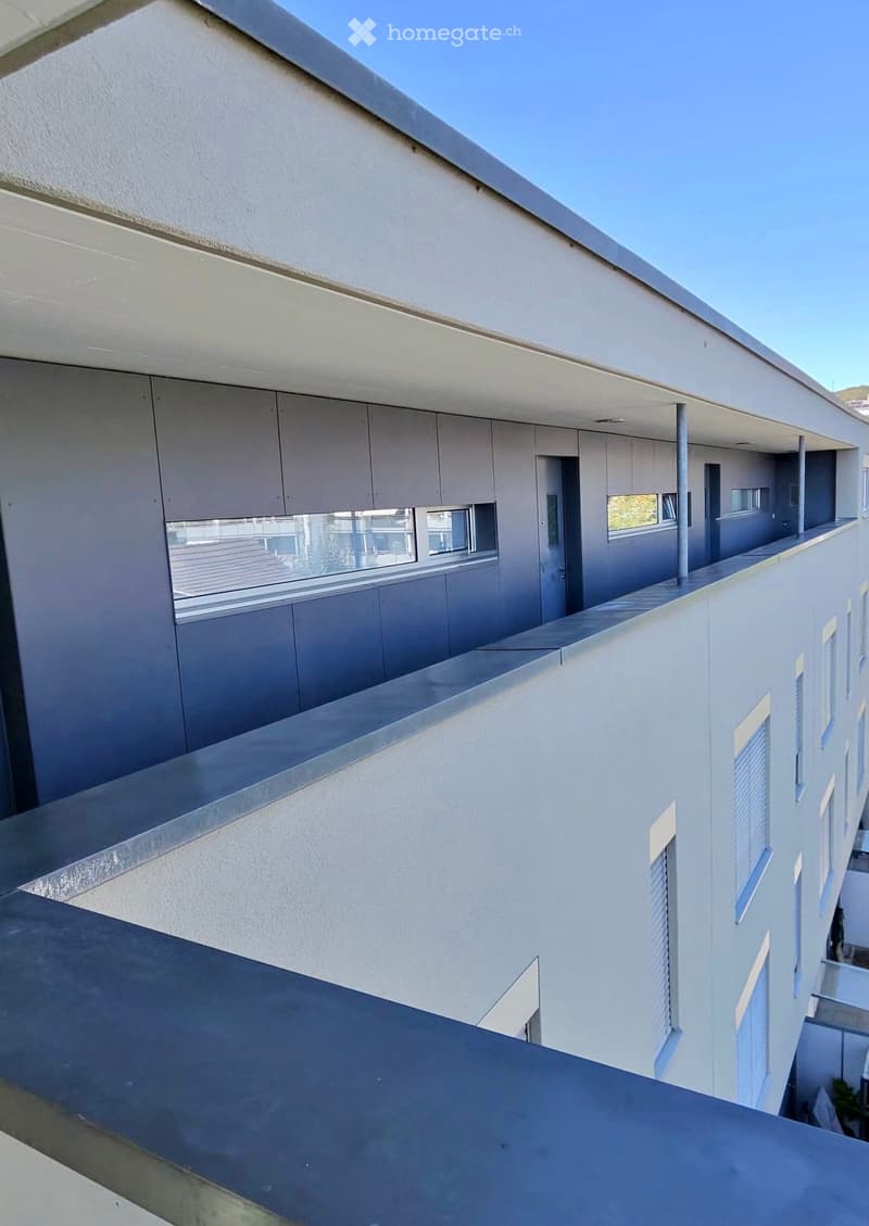Modern balcony with white walls, large windows, and a sleek metal railing, overlooking a clear blue sky.