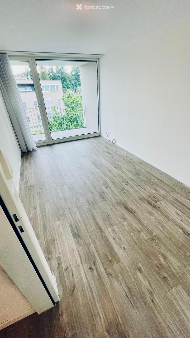 Empty room with light wooden flooring and white walls, featuring a large sliding glass door leading to a balcony with a view of greenery and neighboring buildings.