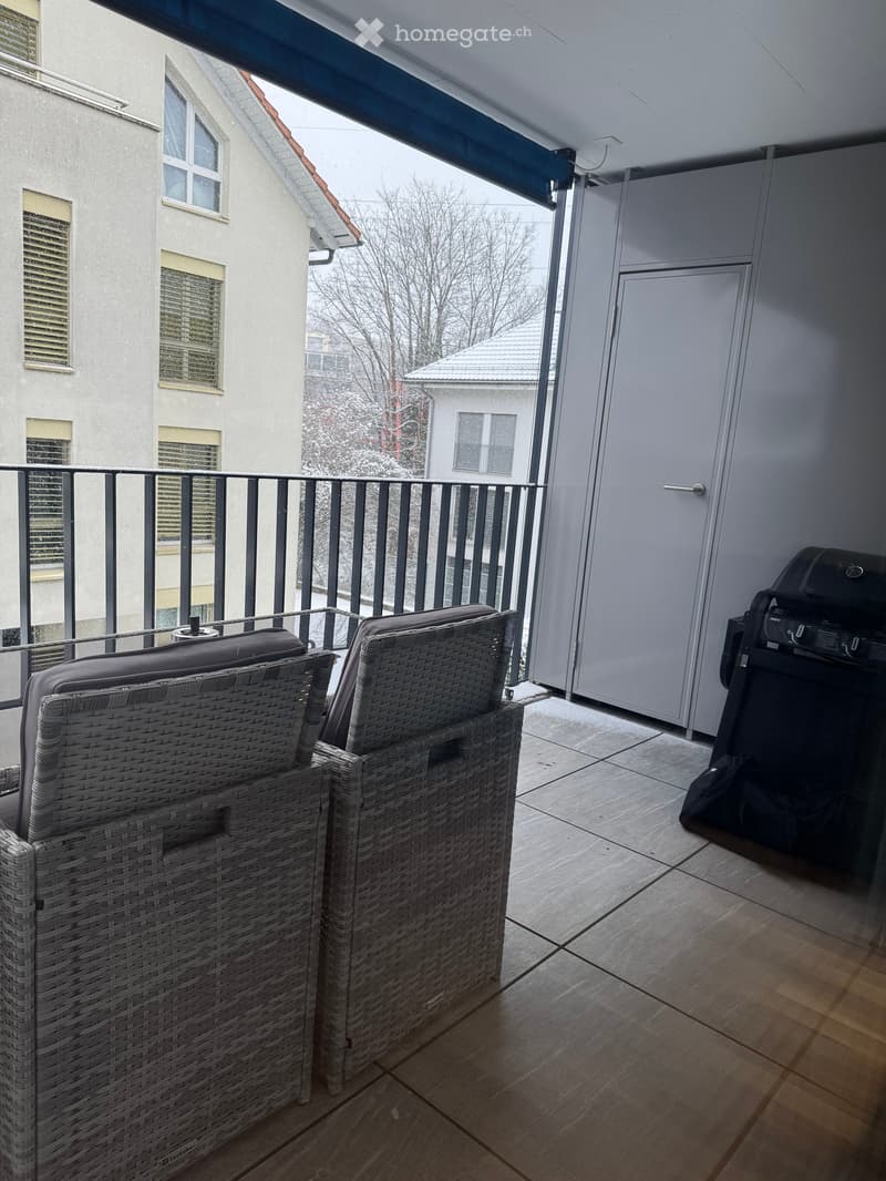 Balcony with two wicker chairs, a grill, and a view of the street and neighboring buildings.