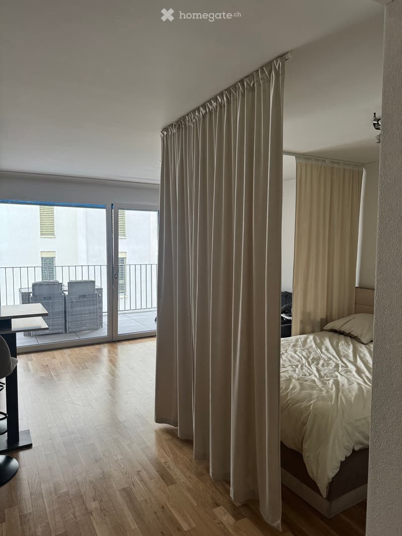 Modern bedroom with wooden flooring, large bed with white bedding, a small table, and a balcony with outdoor seating.