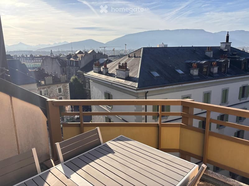 Balcony with wooden railing, table, and chair, overlooking cityscape and mountains.