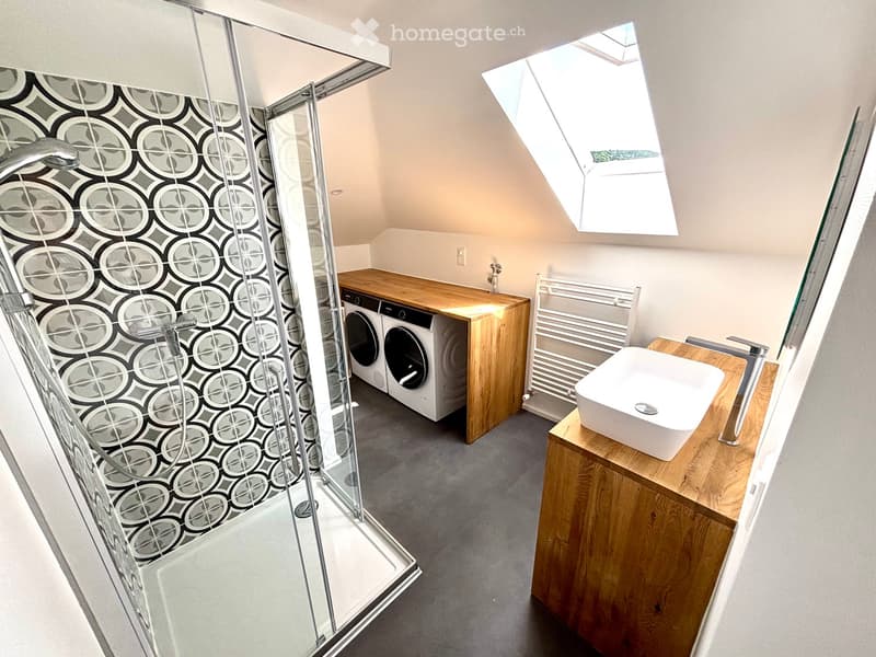 Modern bathroom with a glass-enclosed shower, wooden vanity with sink, washing machine, radiator, and skylight window.