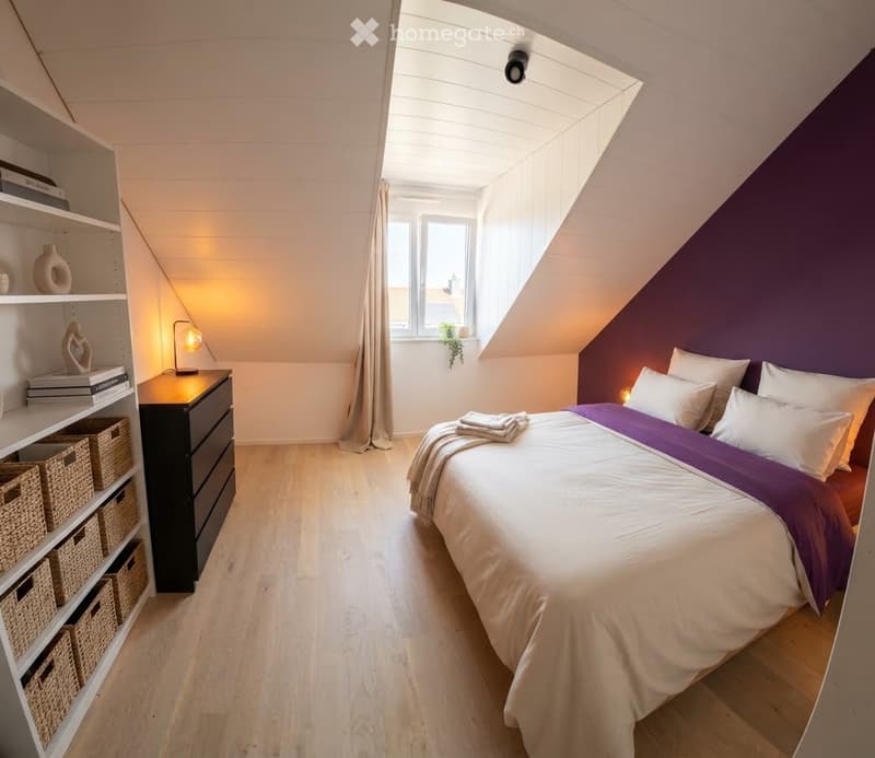 Cozy attic bedroom with slanted ceilings, wooden floors, a large bed with white and purple bedding, a nightstand with a lamp, a built-in shelf with books and decorative items, and wicker baskets on the floor.