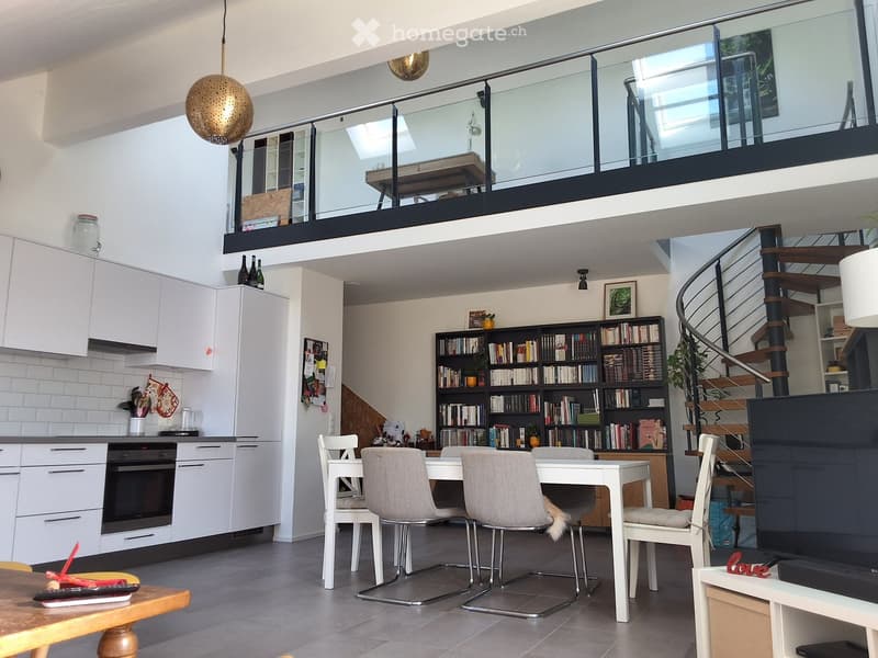 Open-plan living and dining area with modern white cabinets, a dining table with four chairs, a bookshelf, and a staircase leading to a loft area.