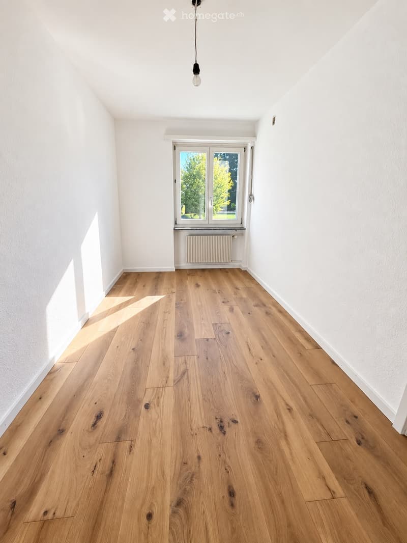 Empty room with light wooden flooring, white walls, and a large window with a radiator below it. Natural light streams in, illuminating the space.