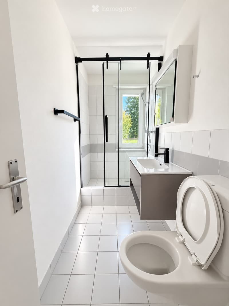 Modern bathroom with white tiles, a toilet, sink, mirror, and a glass-enclosed shower area with a window.
