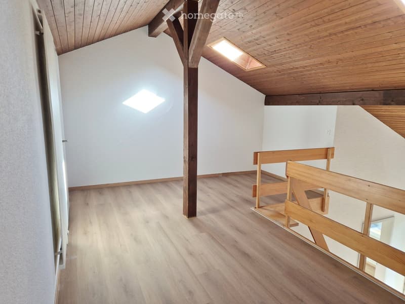 Empty room with wooden flooring, white walls, exposed wooden beams, and a small wooden bench near the corner.