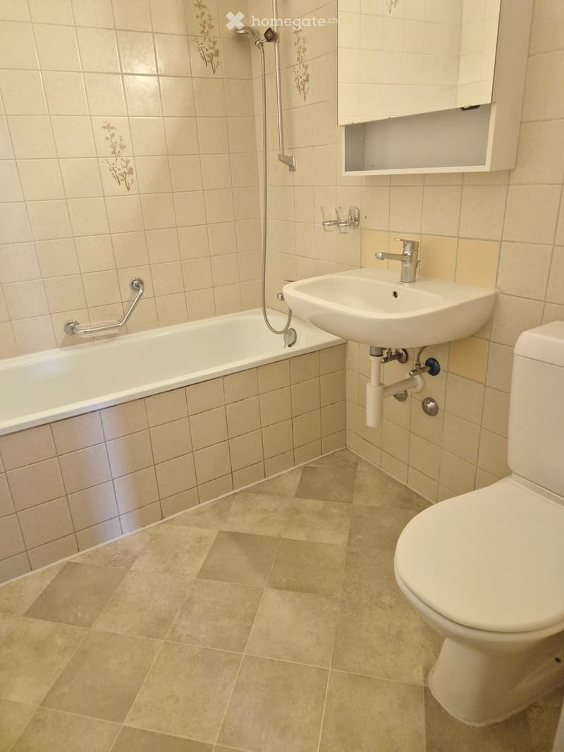 Bathroom with beige tiled walls and floor, featuring a bathtub, sink, toilet, and a small cabinet above the sink.