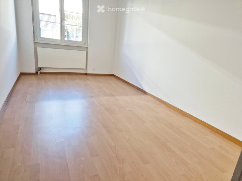 Empty room with light wooden flooring, white walls, and a large window with a radiator below it.