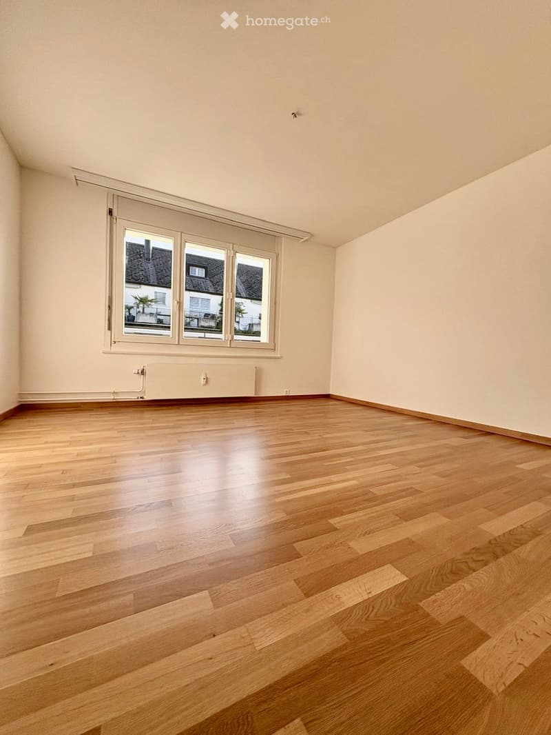Empty room with wooden flooring, large window with white frame, radiator under the window, and light-colored walls.
