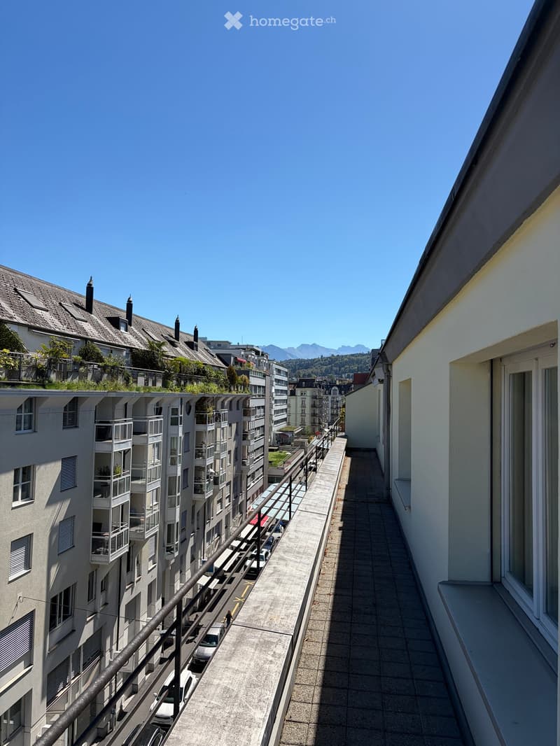Balcony overlooking a city street with parked cars, buildings, and distant mountains under a clear blue sky.