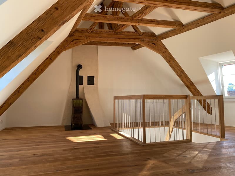Spacious attic room with exposed wooden beams, wooden flooring, skylights, and a staircase railing. A wood-burning stove is visible against the wall.