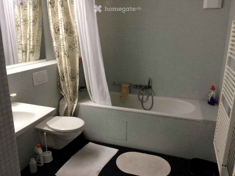 Bathroom with a bathtub, toilet, sink, and shower curtain. Cleaning supplies are placed near the sink and bathtub.