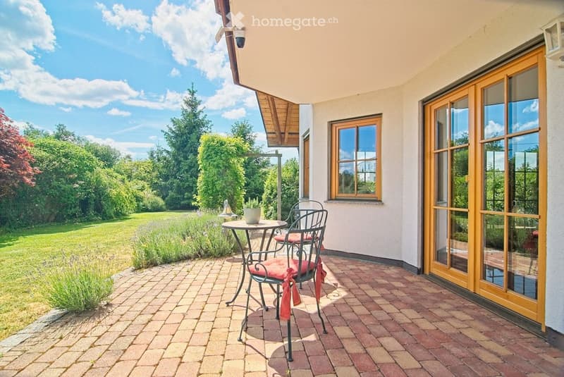Brick-paved balcony with a small round table and two chairs, surrounded by greenery and a view of a lawn.