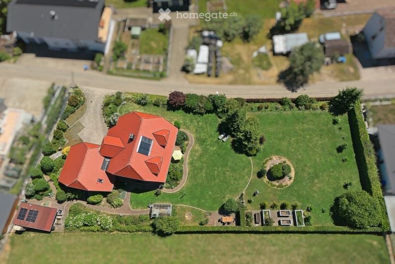 A two-story house with a red-tiled roof, surrounded by a well-maintained garden with pathways, trees, and a circular flower bed.