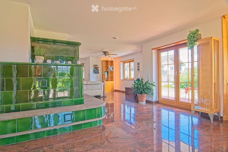 Spacious living room with a green tiled fireplace, white leather sofa, large potted plant, wooden shelf, and glass doors leading to a patio.