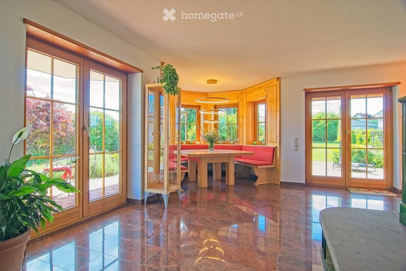 Bright, spacious room with large wooden-framed windows and doors leading to a garden. Features a central wooden table with a red bench seating, a hanging pendant light, and a potted plant near the entrance.
