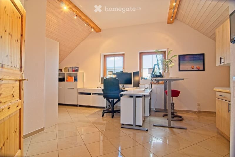 Bright, modern office space with wooden ceiling beams, tiled floor, and large windows. Features a desk with two computer screens, a comfortable chair, a small cabinet, and a potted plant on a side table.