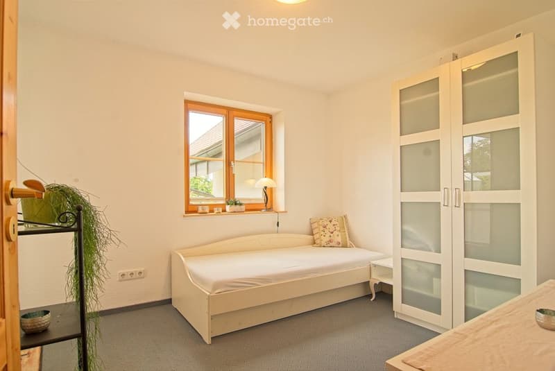 Cozy bedroom with a single bed, white wardrobe with glass panels, small table, lamp on windowsill, and decorative plant near the door.