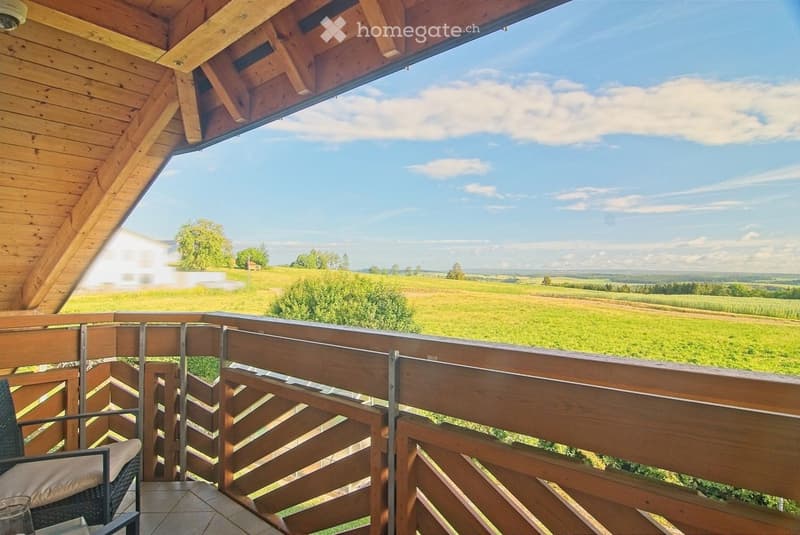 Wooden balcony with metal railing, overlooking a green field and distant landscape under a partly cloudy sky.