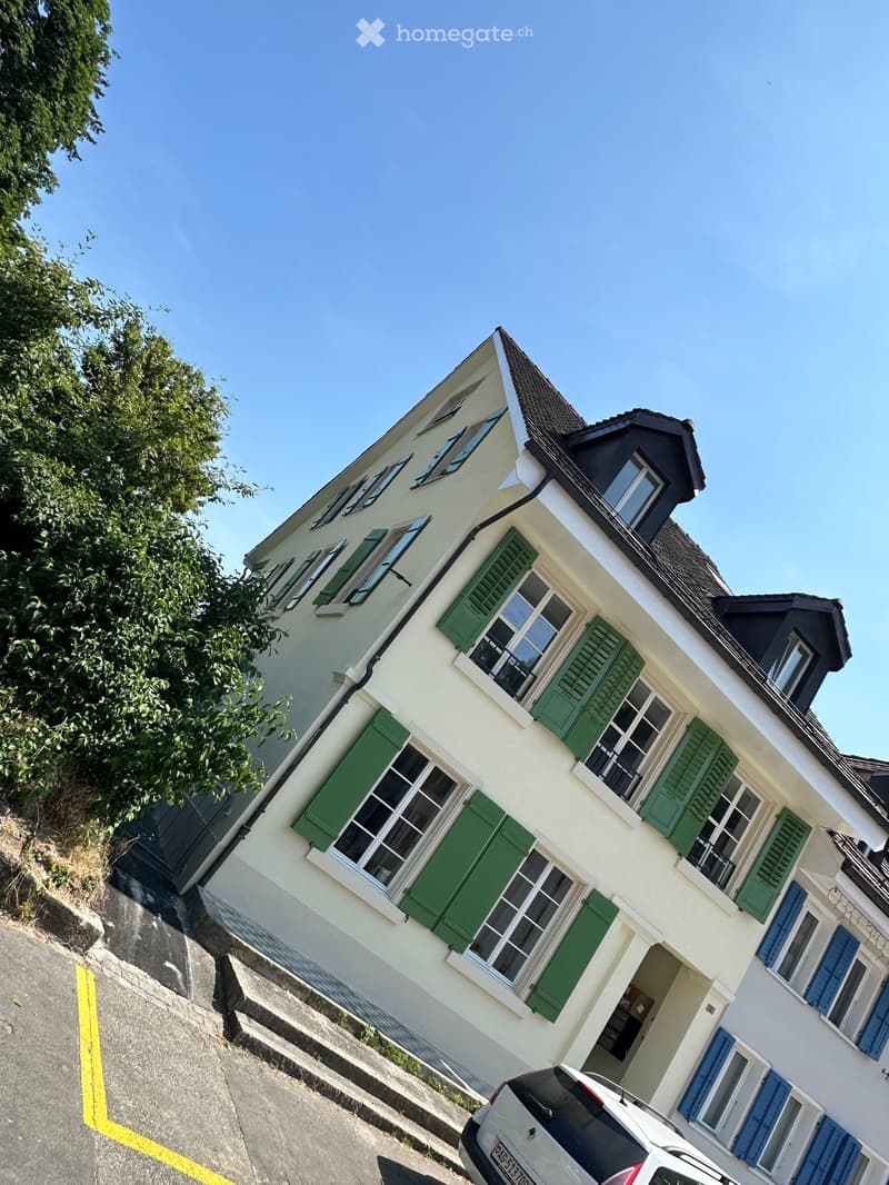 Two-story house with white walls, green shutters, and a dark roof. The house has multiple windows and a visible entrance. A car is parked in front of the house.