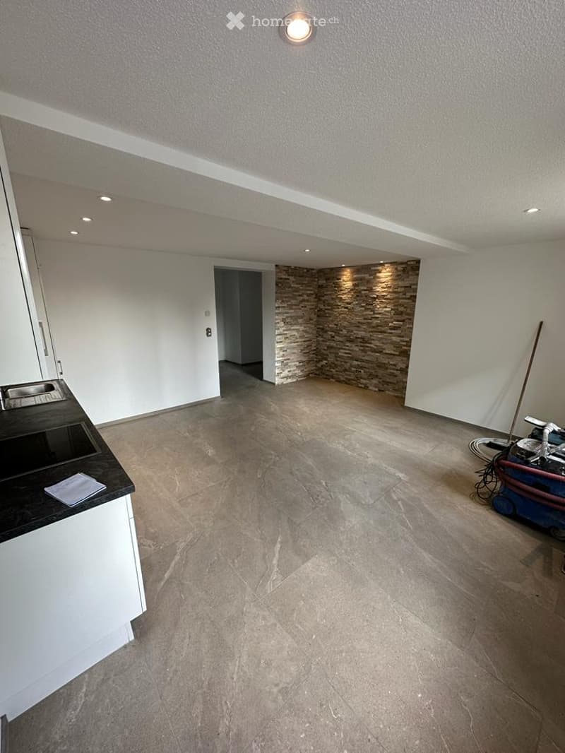 Empty room with white walls, tiled floor, and a small black countertop with a sink. A stone accent wall and a doorway lead to another space.