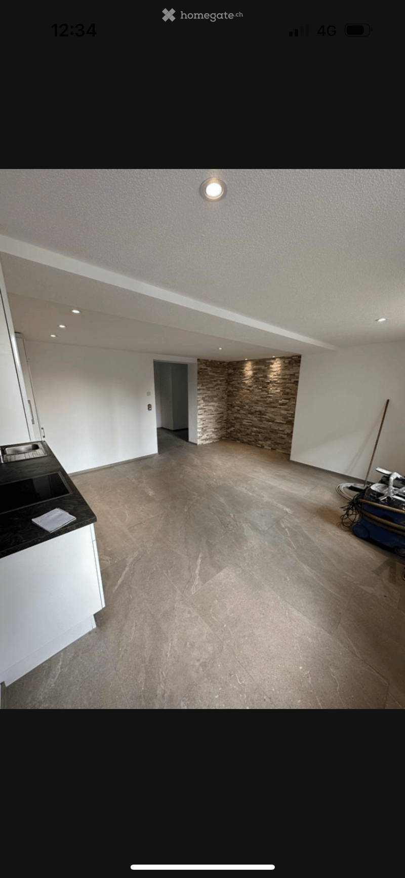 Empty room with white walls, beige tiled floor, and a small black countertop with a sink. A stone accent wall and a doorway lead to another space.