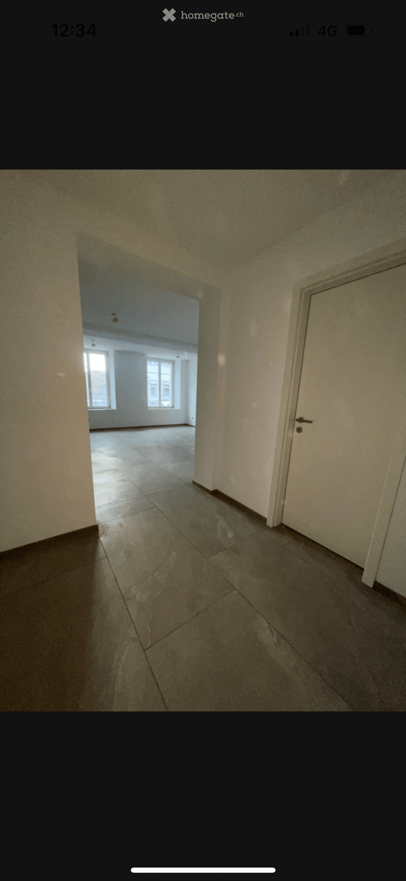 Empty hallway with tiled floor and white walls, leading to another room with windows and a tiled floor.