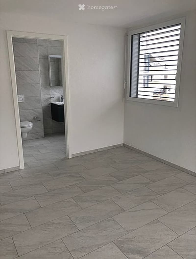 Empty room with grey tiled flooring, white walls, and a doorway leading to a small bathroom with a toilet, sink, and mirror.