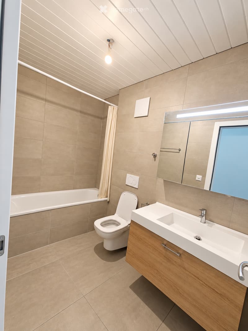 Modern bathroom with beige tiles, a white toilet, a wooden sink cabinet, a bathtub, and a large mirror with built-in lighting.