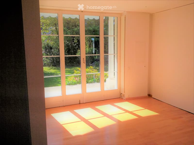 Empty room with wooden flooring and large windows offering a view of a garden. No furniture or equipment is present.