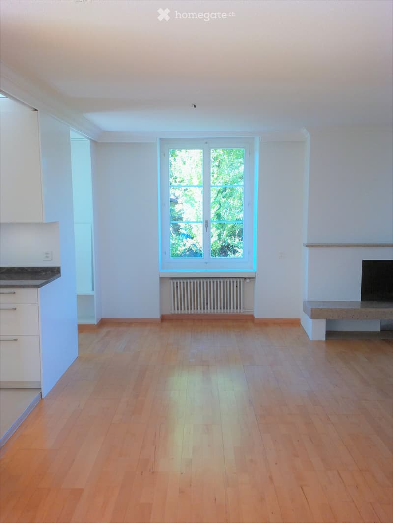 Empty room with light wooden flooring, white walls, and large windows allowing natural light. Features include a radiator, a small bench, and a cabinet with drawers.