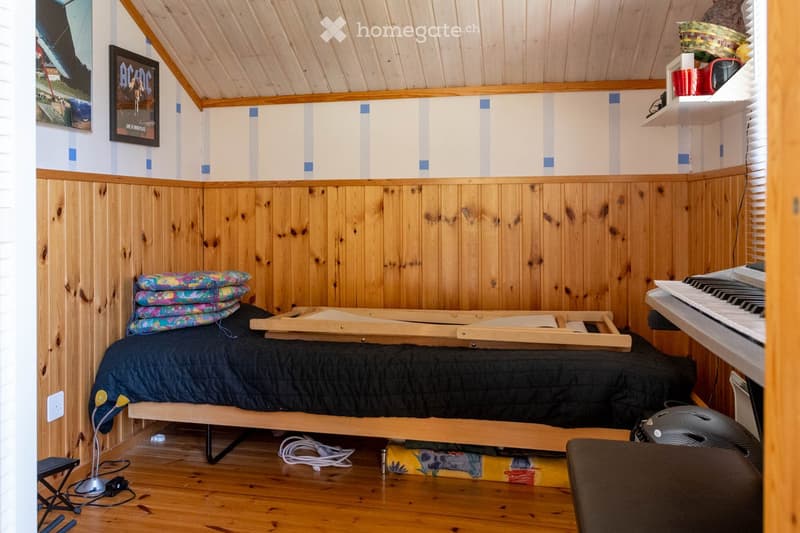Furnished Room. Cozy attic bedroom with wooden walls and ceiling, featuring a bed, small table, piano, shelves, and a picture frame on the wall.