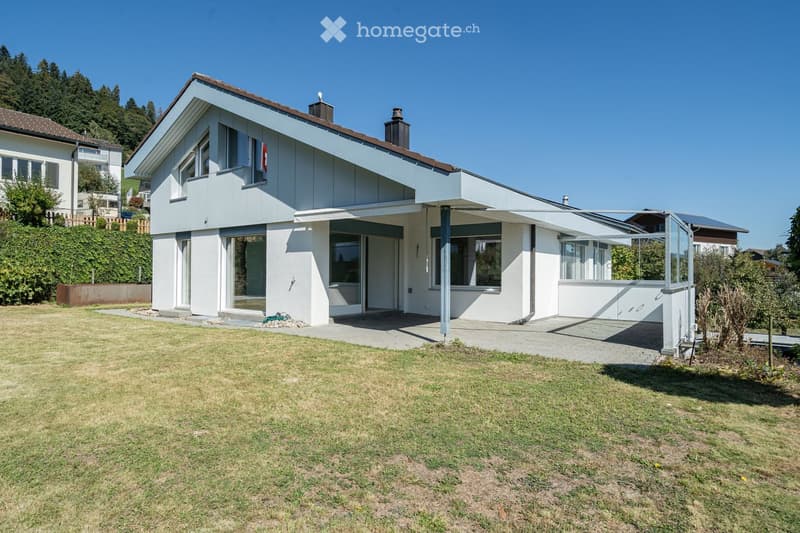 Modern two-story house with white walls, large windows, and a covered patio area. The house features a flat roof, multiple chimneys, and is surrounded by a well-maintained lawn and garden.