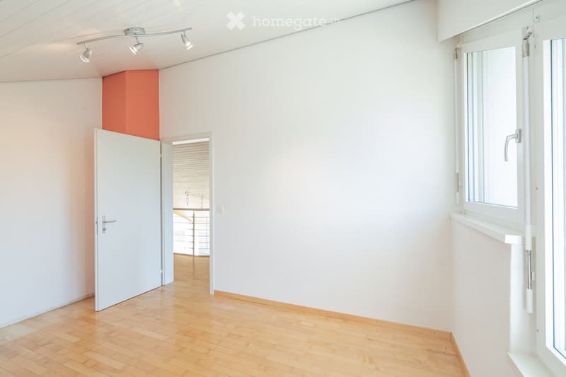 Empty room with white walls, wooden flooring, and a large window with blinds. A door leads to another room with a visible kitchen area.