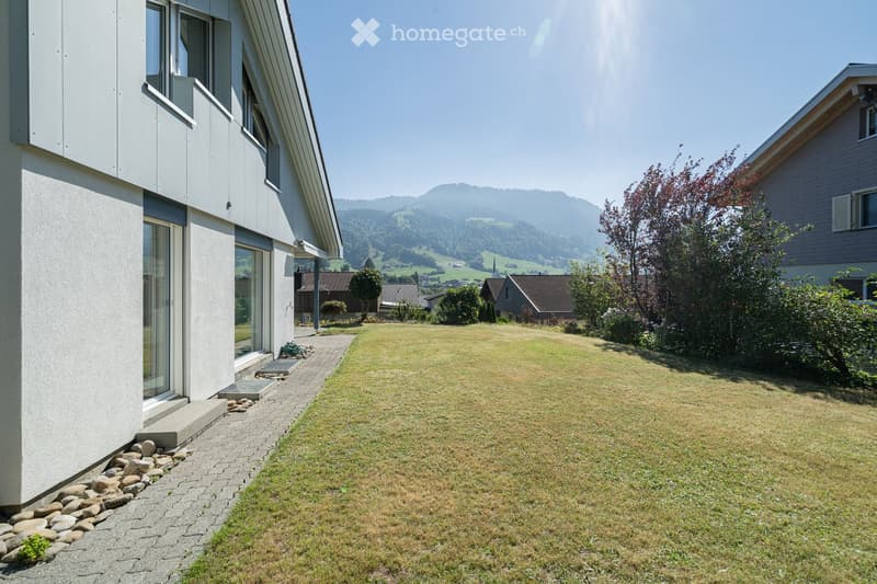 Modern two-story house with white exterior, large windows, and a well-maintained lawn in front. The house features a raised ground floor and a scenic mountain view in the background.
