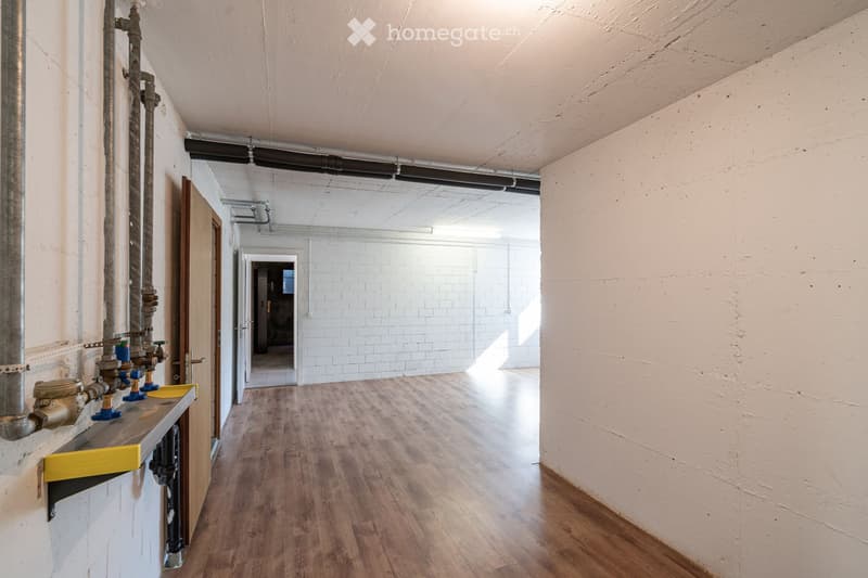 Empty hallway with wooden flooring, white walls, and exposed pipes on one side. Natural light streams in from the far end, revealing an open space beyond.