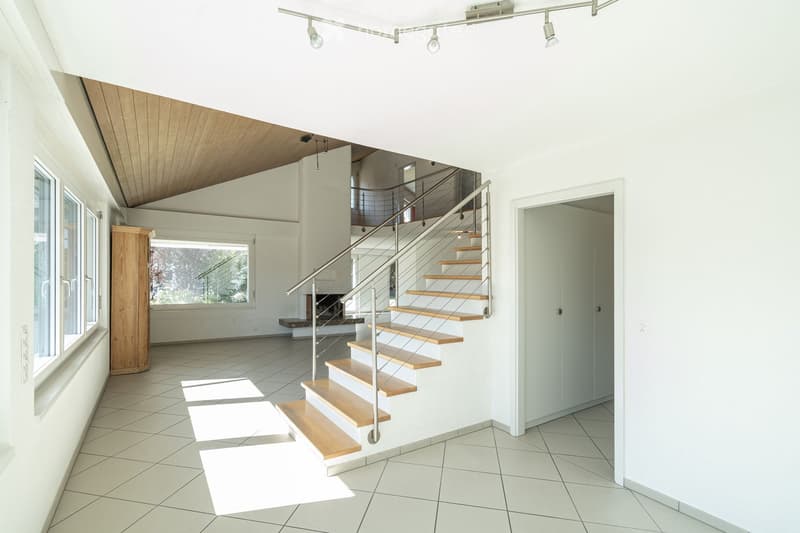Modern hallway with a wooden staircase, white walls, and large windows allowing natural light. The ceiling features recessed lighting and a wooden slanted section.
