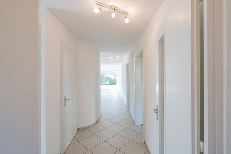 A clean, modern hallway with white tiled flooring, white walls, and recessed lighting. Doors are visible on both sides, leading to other rooms.