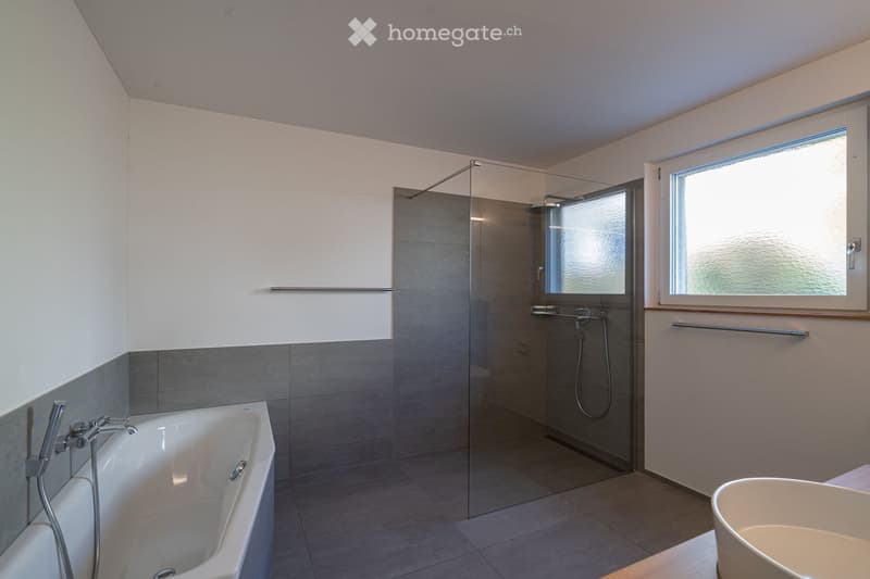 Modern bathroom with a bathtub, glass-enclosed shower, and large window providing natural light. Features grey tiles, white walls, and chrome fixtures.