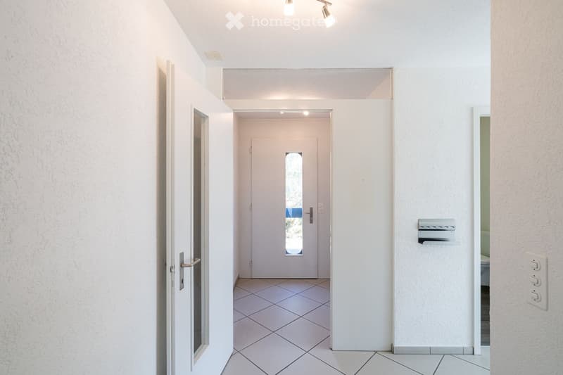 A clean, modern hallway with white walls, tiled flooring, and recessed lighting. Features include a white door with a small window, a mail slot, and a light switch on the wall.