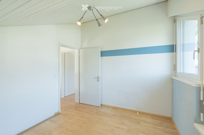 Empty room with light-colored walls, wooden flooring, and a ceiling light fixture. There is a window with a blue accent strip and a door leading to another room.