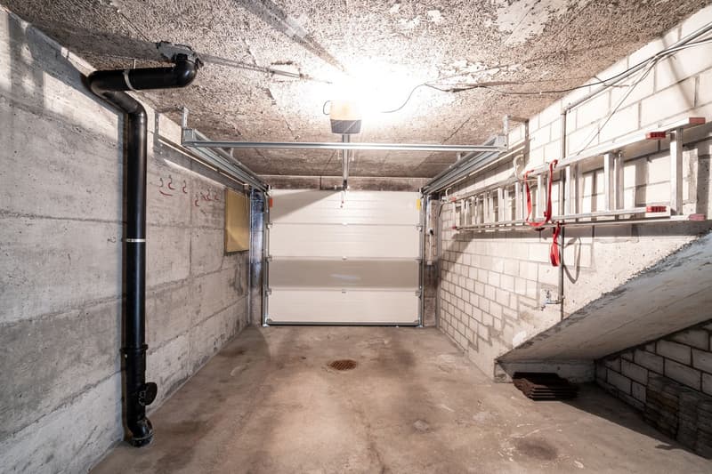 Empty concrete-walled corridor with a garage door at the end, metal shelving, and exposed pipes.