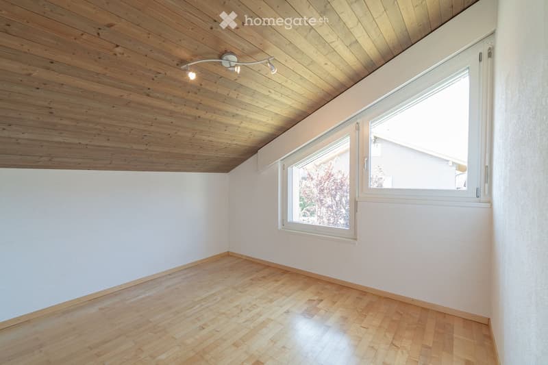 Empty room with wooden flooring, white walls, and a large window with white frame. The ceiling is wooden with built-in lighting.