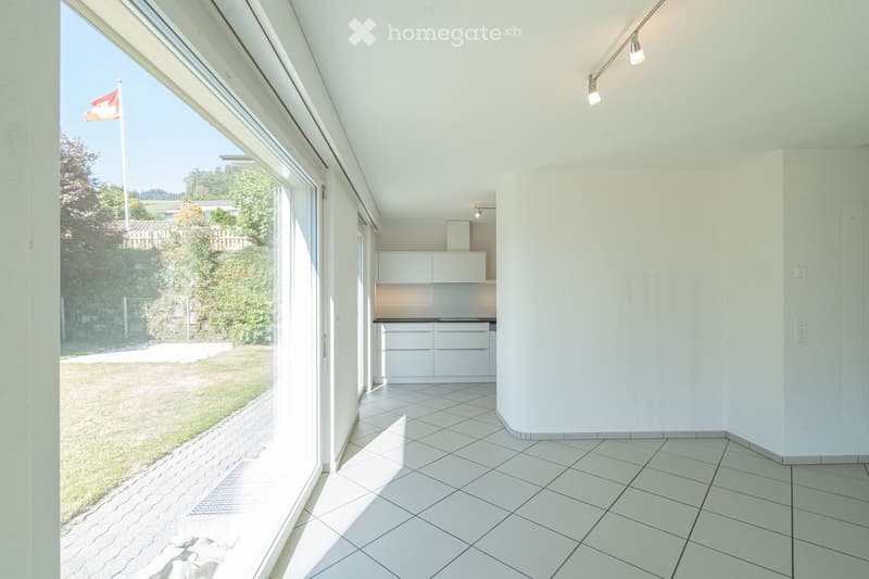 Bright, unfurnished room with white tiled flooring, white walls, and large windows offering a view of a green outdoor area. The room features modern lighting fixtures and a small, white kitchen area with cabinets and a countertop.