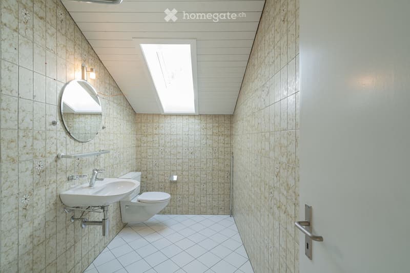 Modern bathroom with beige tiled walls, white tiled floor, a sink, toilet, and a round mirror above the sink. The room features a skylight window and a light fixture on the wall.