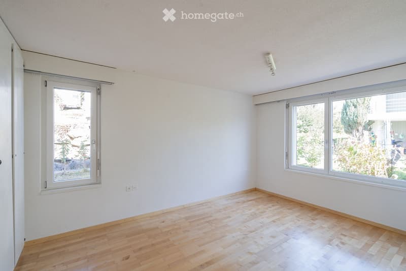 Empty room with wooden flooring, white walls, and large windows offering views of greenery and a street.