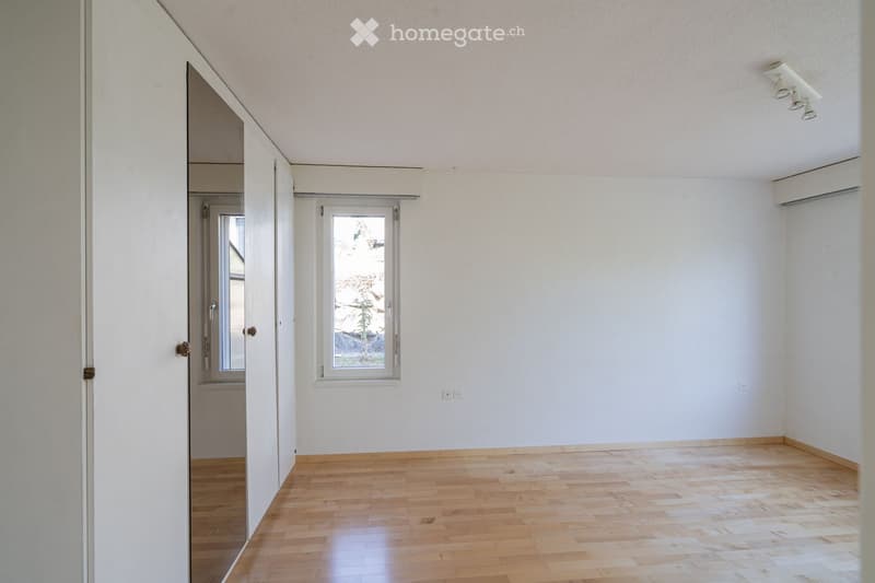 Empty room with light wooden flooring, white walls, and large windows allowing natural light. The room features a sliding door leading to another space and a visible air conditioning unit on the wall.