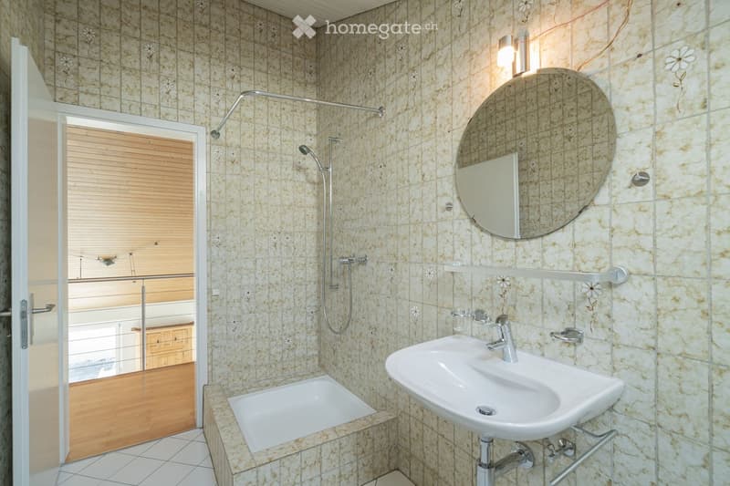 Bathroom with beige tiled walls, a white sink, a bathtub, a large round mirror, and a shower with a handheld sprayer. The room features a wooden door with blinds and a small shelf above the sink.